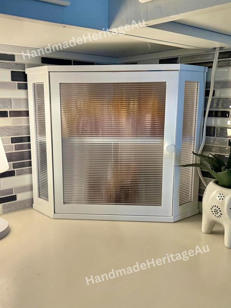 May include: White corner cabinet with a ribbed glass door and side panels. The cabinet has a small handle and is mounted on a countertop. The text "HandmadeHeritageAu" is visible on the top and bottom of the cabinet.