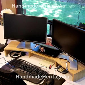 May include: A home office setup with two computer monitors on a light wood monitor stand. A black keyboard, mouse, and headphones are visible. A laptop and printer are also on the desk. The text "HandmadeHeritageAu" is visible.