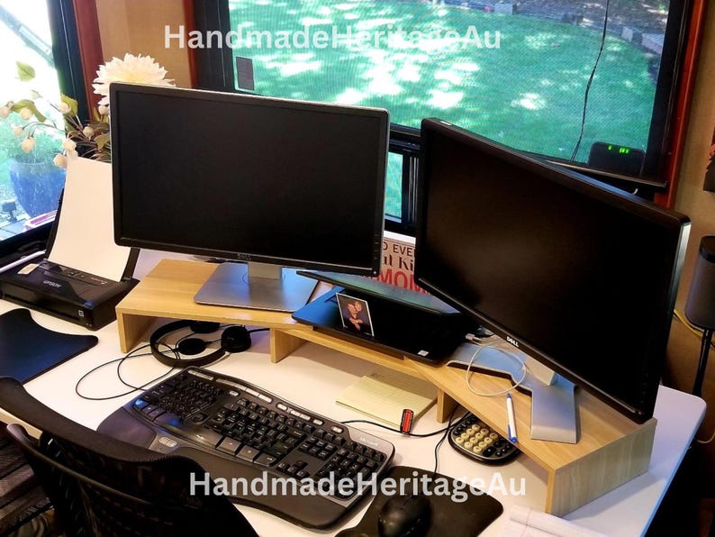 May include: A home office setup with two computer monitors on a light wood monitor stand. A black keyboard, mouse, and headphones are visible. A laptop and printer are also on the desk. The text "HandmadeHeritageAu" is visible.