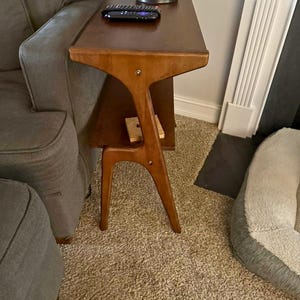 K&ouml;nnte beinhalten: Ein Beistelltisch im Mid-Century-Modern-Stil in warmbraunem Finish. Der Tisch hat ein einzigartiges, skulpturales Design mit einer dreieckigen Oberseite, einem unteren Regal und abgewinkelten Beinen. Eine Fernbedienung liegt auf der Tischoberfl&auml;che.