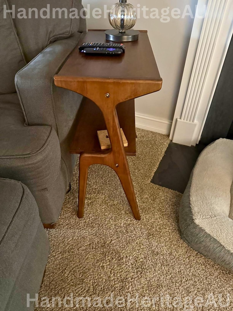 K&ouml;nnte beinhalten: Ein Beistelltisch im Mid-Century-Modern-Stil in warmbraunem Finish. Der Tisch hat ein einzigartiges, skulpturales Design mit einer dreieckigen Oberseite, einem unteren Regal und abgewinkelten Beinen. Eine Fernbedienung liegt auf der Tischoberfl&auml;che.