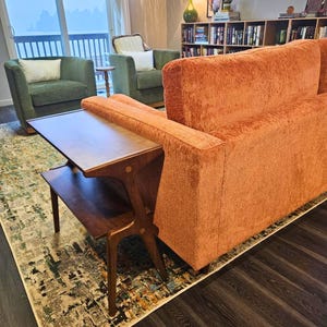 K&ouml;nnte beinhalten: Ein Wohnzimmer im Mid-Century-Modern-Stil mit einem orangefarbenen Sofa, zwei gr&uuml;nen Sesseln und einem Beistelltisch aus Holz. Ein gemusterter Teppich und dunkler Holzboden vervollst&auml;ndigen den Raum. Der Raum ist gut beleuchtet.