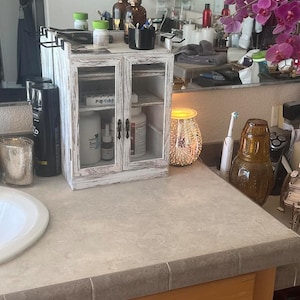 May include: A small, white-washed wooden cabinet with glass doors sits on a bathroom countertop. The cabinet has two doors and a distressed finish. Various toiletries and personal care items are visible inside and on top of the cabinet. A decorative light and a vase of purple orchids are also in the scene.