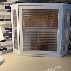 May include: White corner cabinet with a ribbed glass door and side panels. The cabinet has a simple, modern design and is mounted on a light-colored countertop. The text "HandmadeHeritageAu" is visible on the top and bottom of the cabinet.