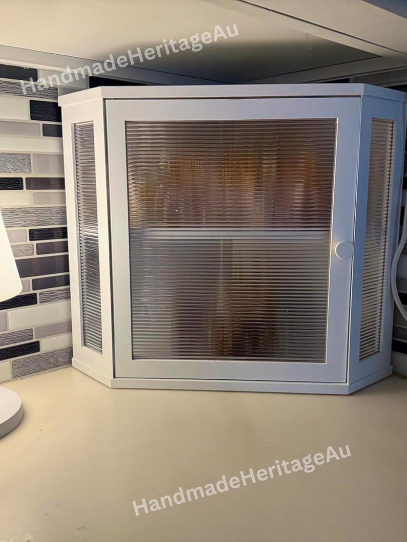 May include: White corner cabinet with a ribbed glass door and side panels. The cabinet has a simple, modern design and is mounted on a light-colored countertop. The text "HandmadeHeritageAu" is visible on the top and bottom of the cabinet.