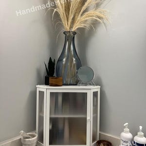 May include: A white corner cabinet with ribbed glass doors and a small shelf. The cabinet is topped with a blue vase holding dried pampas grass. A small mirror and decorative items sit on top of the cabinet.
