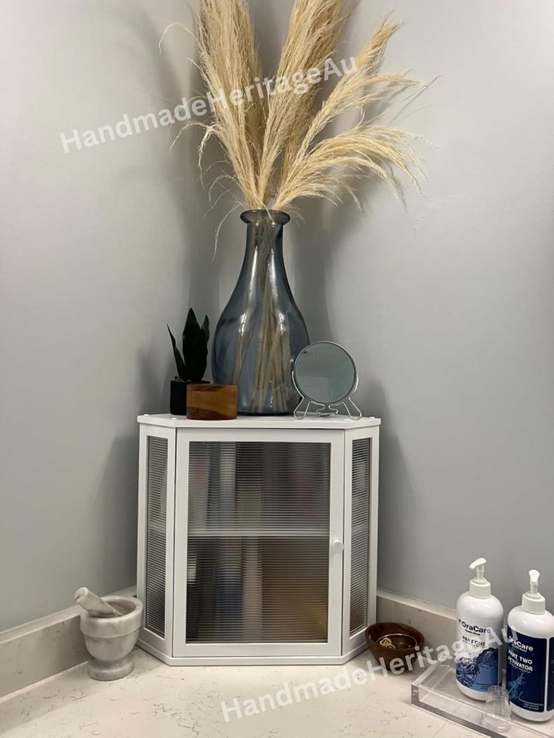 May include: A white corner cabinet with ribbed glass doors and a small shelf. The cabinet is topped with a blue vase holding dried pampas grass. A small mirror and decorative items sit on top of the cabinet.
