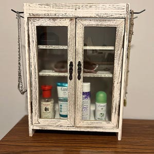 May include: A small, distressed white wooden cabinet with glass doors. The cabinet has black metal hardware and is filled with various toiletries. Jewelry hangs on hooks on either side of the cabinet.
