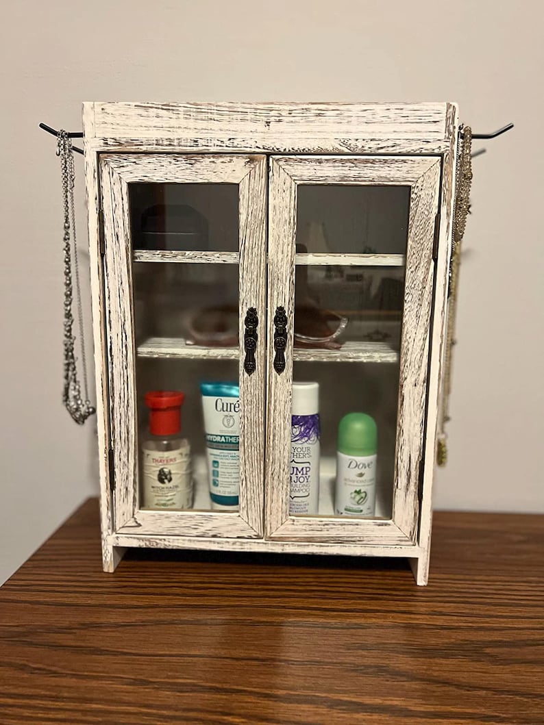 May include: A small, distressed white wooden cabinet with glass doors. The cabinet has black metal hardware and is filled with various toiletries. Jewelry hangs on hooks on either side of the cabinet.