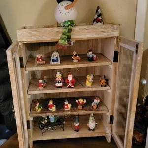 May include: A wooden cabinet with multiple shelves displaying various Christmas ornaments. The ornaments include a snowman, Santa Claus figures, angels, and other festive decorations. The cabinet has glass-paneled doors and is made of light-colored wood.