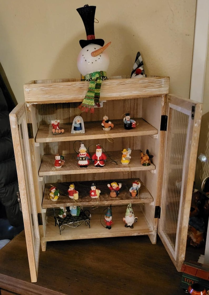 May include: A wooden cabinet with multiple shelves displaying various Christmas ornaments. The ornaments include a snowman, Santa Claus figures, angels, and other festive decorations. The cabinet has glass-paneled doors and is made of light-colored wood.
