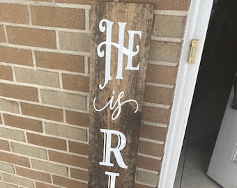 May include: A wooden sign with the words "He is Risen" painted in white on a brown background. The sign is leaning against a brick wall.
