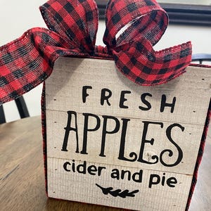 May include: A wooden box with the words "FRESH APPLES cider and pie" in black lettering. The box is decorated with a red and black plaid ribbon bow.