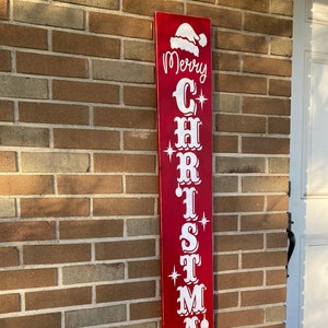 Op de afbeelding: Een rood houten bord met witte letters waarop "Merry Christmas" staat met een kerstmanmuts bovenop.