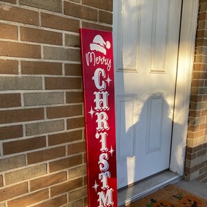 Op de afbeelding: Een rood houten bord met witte letters waarop "Merry Christmas" staat, met een kerstmanmuts bovenop.