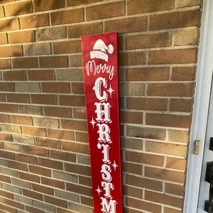 Op de afbeelding: Een rood houten bord met witte letters waarop "Merry Christmas" staat, met een kerstmanmuts bovenop.