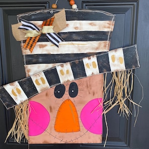 May include: A decorative door hanger shaped like a scarecrow wearing a top hat with black and white stripes. The hat features a burlap, orange plaid, and black and white striped bow. The face has black eyes, a bright orange nose, and pink cheeks.