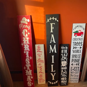 Op de afbeelding: Vijf houten borden met geschilderde kerst- en familiethema's. De borden zijn geschilderd in rood, wit, zwart en cr&egrave;me. De borden tonen de opschriften "Merry Christmas", "and to all a good night", "FAMILY", "Warm Up at the Hot Chocolate Bar Marshmallows" en "Farm Fresh Christmas Trees Noble Spruce Pine U-Cut We-Cut Hot Cocoa Old Fashioned Family Fun".