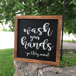 May include: A wooden framed sign with a black chalkboard background. The sign reads "Wash your hands - ya filthy animal -" in white lettering.