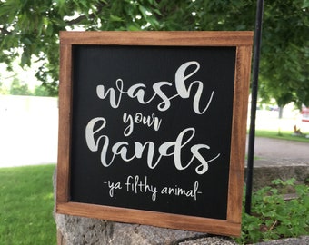 May include: A wooden framed sign with a black chalkboard background. The sign reads "Wash your hands - ya filthy animal -" in white lettering.
