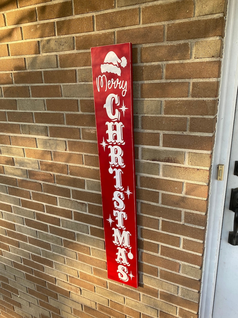 Op de afbeelding: Een rood houten bord met witte letters waarop "Merry Christmas" staat, met een kerstmanmuts bovenop.