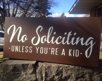 May include: A brown wooden sign with white text that reads "No Soliciting - Unless You're A Kid."