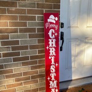 Op de afbeelding: Een rood houten bord met witte letters waarop "Merry Christmas" staat met een kerstmanmuts bovenop.