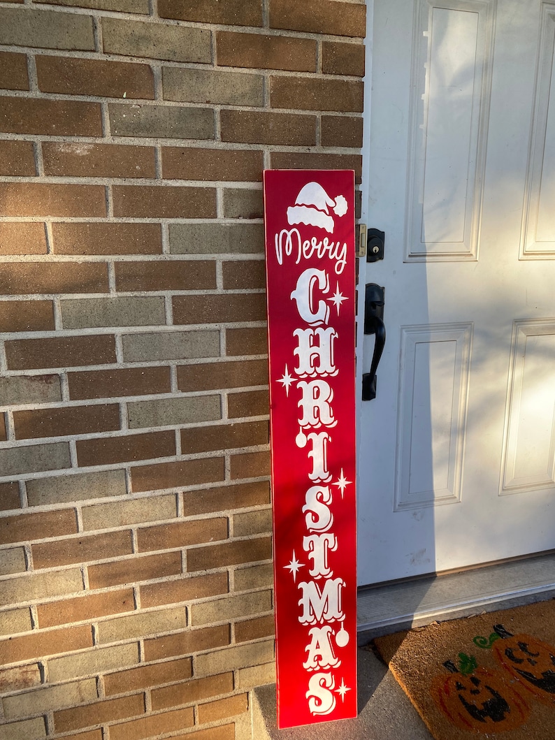 Op de afbeelding: Een rood houten bord met witte letters waarop "Merry Christmas" staat met een kerstmanmuts bovenop.