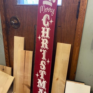 Op de afbeelding: Een rood houten bord met witte letters waarop "Merry Christmas" staat. Het bord heeft bovenaan een kerstmanmuts en sterren in de letters.