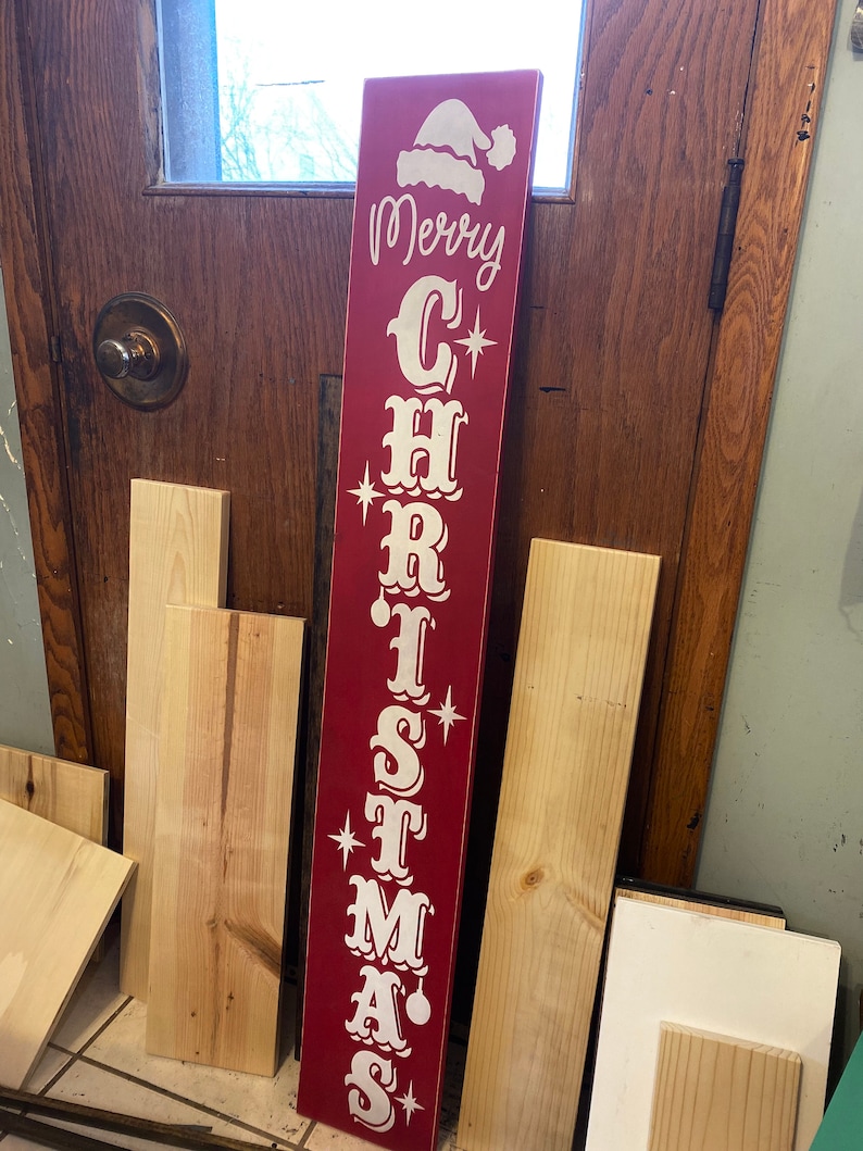 Op de afbeelding: Een rood houten bord met witte letters waarop "Merry Christmas" staat. Het bord heeft bovenaan een kerstmanmuts en sterren in de letters.