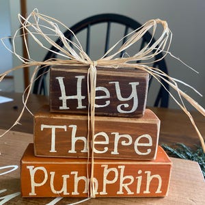 May include: A stack of three wooden blocks with the words "Hey There Pumpkin" painted in white on them. The blocks are tied together with a brown raffia bow.