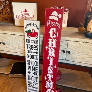 Op de afbeelding: Twee houten kerstversieringen. De versiering links is wit met zwarte tekst en zegt "Farm Fresh Christmas Trees Noble Spruce Pine U-Cut We-Cut Hot Cocoa Old Fashioned Family Fun". De versiering rechts is rood met witte tekst en zegt "Merry Christmas".