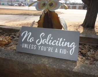 May include: A gray wooden sign with white text that reads "No Soliciting - Unless You're A Kid."