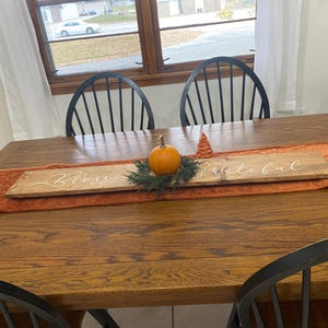 May include: A wooden table with a brown table runner and a wooden sign that says "Bless & Be Thankful". The sign is decorated with a small pumpkin and a pine sprig.