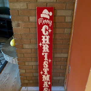 Op de afbeelding: Een rood houten bord met witte letters waarop "Merry Christmas" staat, met een kerstmanmuts bovenop.