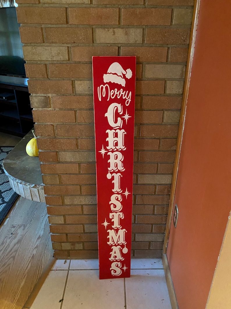 Op de afbeelding: Een rood houten bord met witte letters waarop "Merry Christmas" staat, met een kerstmanmuts bovenop.