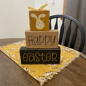 May include: A three-tiered wooden block sign with the words "Happy Easter" painted in black. The top block features a painted bunny with a yellow ribbon.