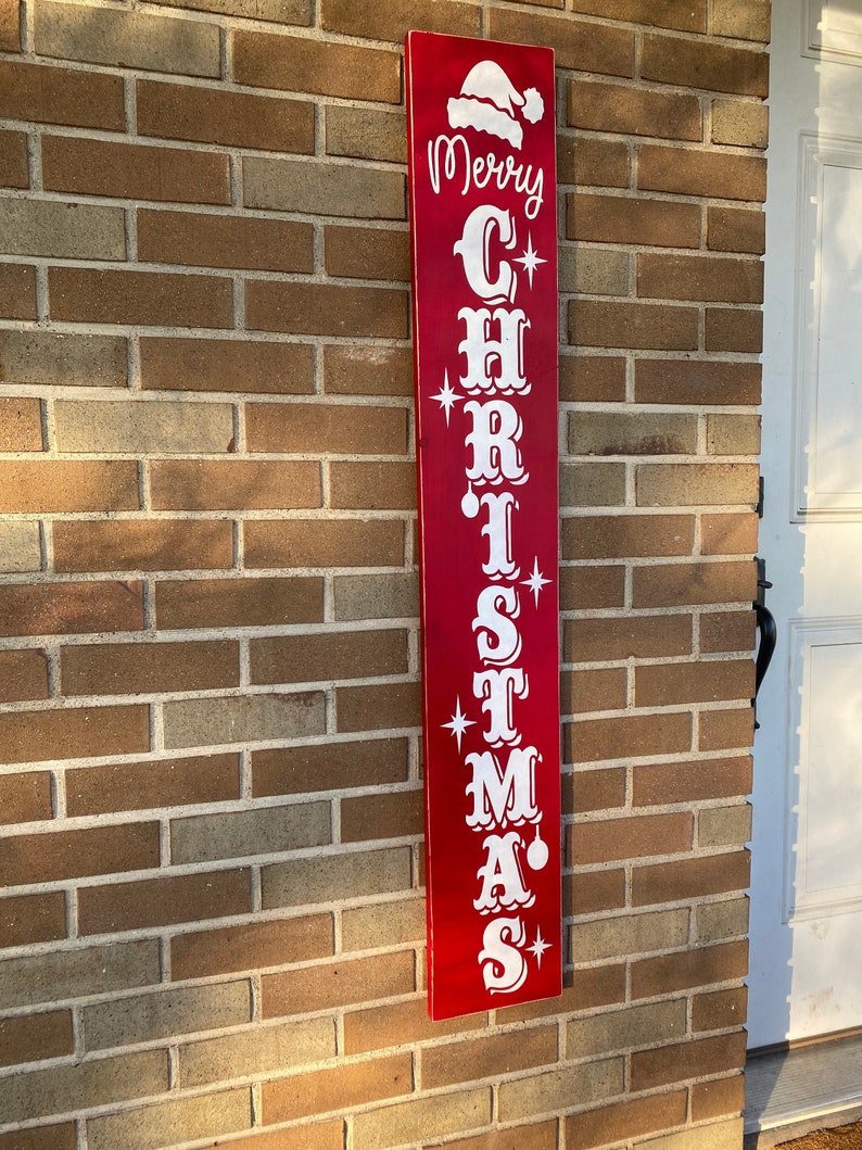 Op de afbeelding: Een rood houten bord met witte letters waarop "Merry Christmas" staat met een kerstmanmuts bovenop.