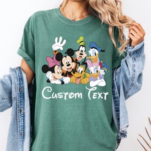 May include: Seafoam green t-shirt with a graphic of Disney characters: Mickey Mouse, Minnie Mouse, Donald Duck, Daisy Duck, Goofy, and Pluto. The words "Custom Text" are written in white script across the bottom of the graphic.