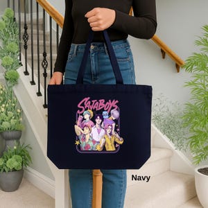 May include: Navy blue tote bag featuring a colorful graphic of six stylized figures with the text "SataBoys" in a pink outlined font. The bag is held by a person. The word "Navy" is printed at the bottom right.