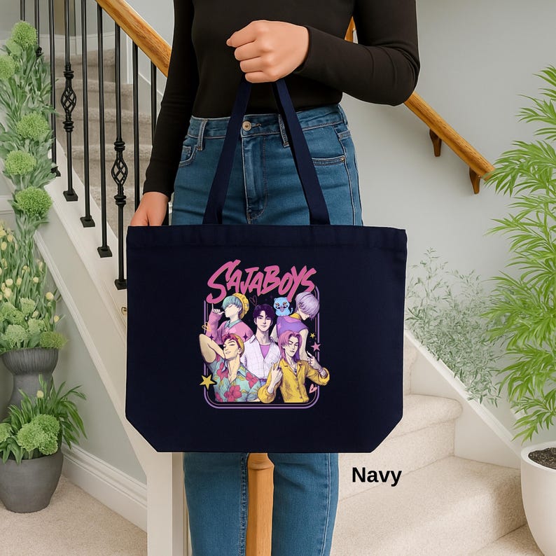 May include: Navy blue tote bag featuring a colorful graphic of six stylized figures with the text "SataBoys" in a pink outlined font. The bag is held by a person. The word "Navy" is printed at the bottom right.
