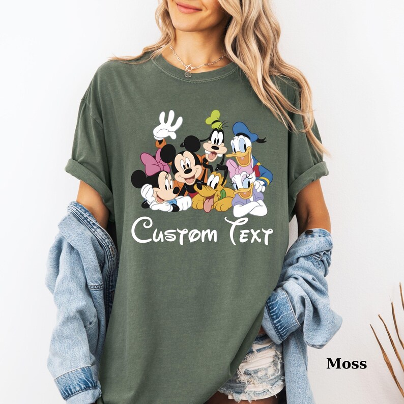 May include: A moss green t-shirt with a cartoon graphic of Mickey and Minnie Mouse, Goofy, Donald and Daisy Duck, and Pluto. The words "Custom Text" are written in white script below the characters. The shirt has a relaxed fit.