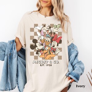 May include: Ivory-colored t-shirt featuring a vintage-style graphic of Mickey Mouse and friends. The design includes Mickey, Minnie, Donald, Daisy, Goofy, and Pluto, set against a checkered background. The text "MICKEY & CO. EST. 1928" is printed below the characters.