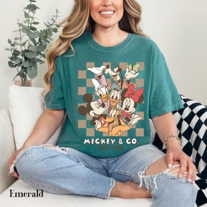 May include: Emerald green t-shirt featuring a graphic of Mickey Mouse and friends. The design includes Mickey, Minnie, Donald, Daisy, Goofy, and Pluto, with the text "Mickey & Co. Est. 1928" on a checkered background.
