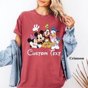 May include: Crimson t-shirt with a graphic of Disney characters, including Mickey and Minnie Mouse, Goofy, Donald Duck, Daisy Duck, and Pluto. The words "Custom Text" are printed in white script. The shirt has short sleeves.