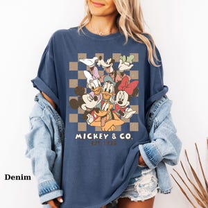 May include: Denim blue t-shirt featuring a graphic of Mickey Mouse and friends. The design includes Mickey, Minnie, Donald Duck, Daisy Duck, Goofy, and Pluto, set against a checkered background. The text "MICKEY & CO. EST 1928" is printed below the characters.
