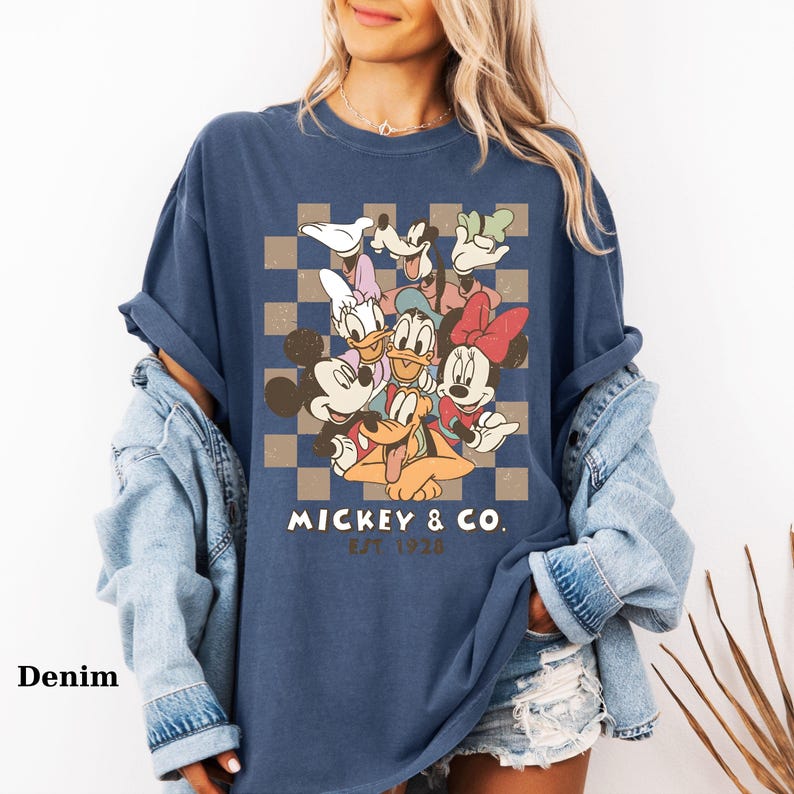May include: Denim blue t-shirt featuring a graphic of Mickey Mouse and friends. The design includes Mickey, Minnie, Donald Duck, Daisy Duck, Goofy, and Pluto, set against a checkered background. The text "MICKEY & CO. EST 1928" is printed below the characters.