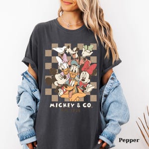 May include: Dark gray t-shirt featuring a colorful graphic of Disney characters, including Mickey Mouse, Minnie Mouse, Donald Duck, and Goofy, set against a checkered background. The text "Mickey & Co. Est. 1928" is printed below the graphic.