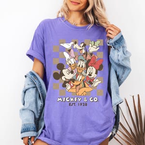 May include: A purple t-shirt featuring a graphic of Mickey Mouse, Minnie Mouse, Donald Duck, Daisy Duck, Goofy, and Pluto. The design includes a brown and tan checkered background and the text "Mickey & Co. Est. 1928".