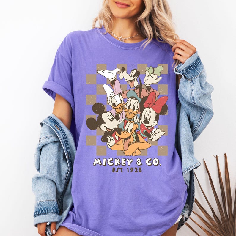 May include: A purple t-shirt featuring a graphic of Mickey Mouse, Minnie Mouse, Donald Duck, Daisy Duck, Goofy, and Pluto. The design includes a brown and tan checkered background and the text "Mickey & Co. Est. 1928".